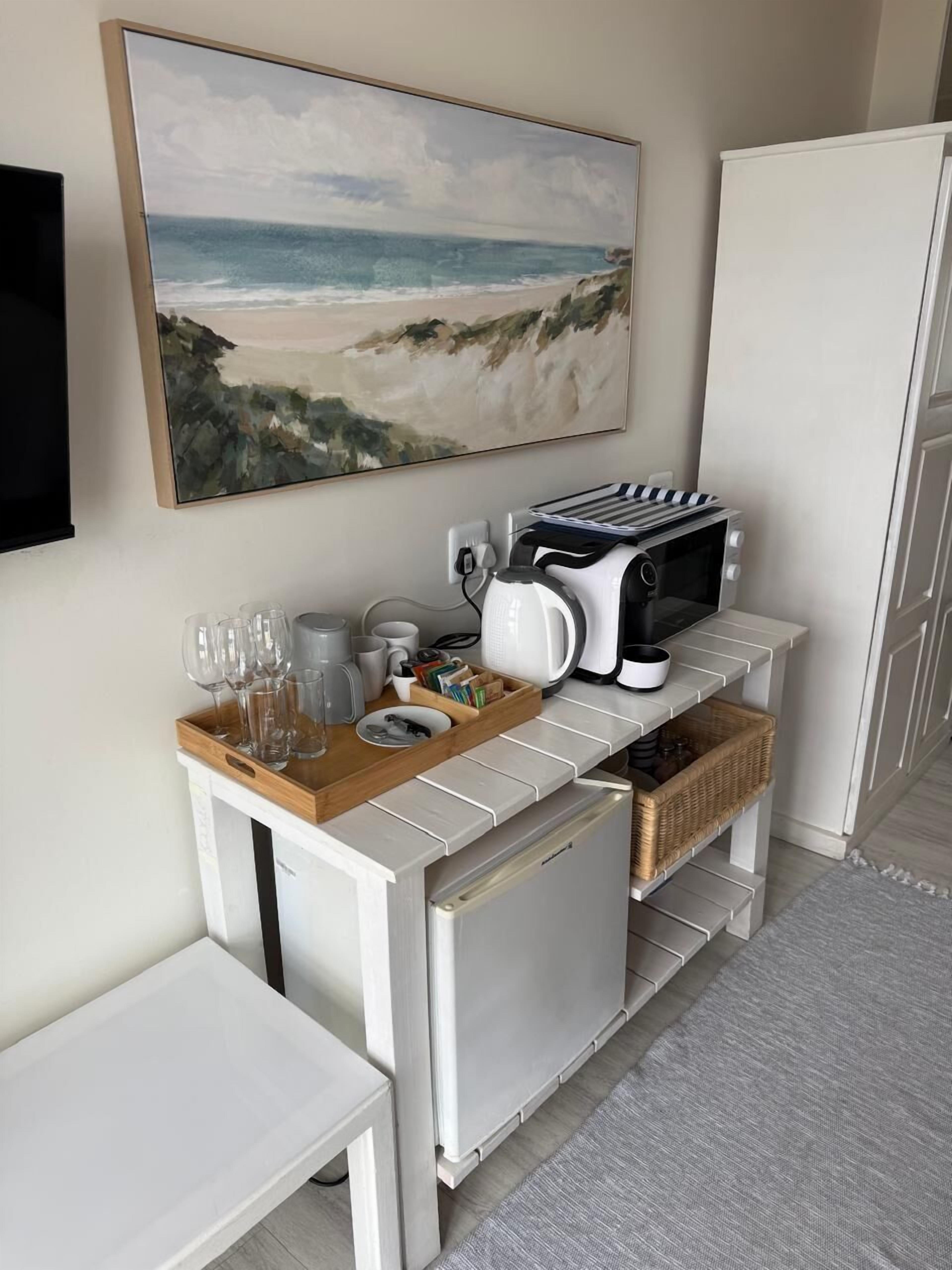 Un angolo cottura con forno a microonde, tostapane e macchina del caffè su un piano di lavoro bianco. Calici da vino e tazze su un vassoio. Un quadro raffigura una spiaggia.