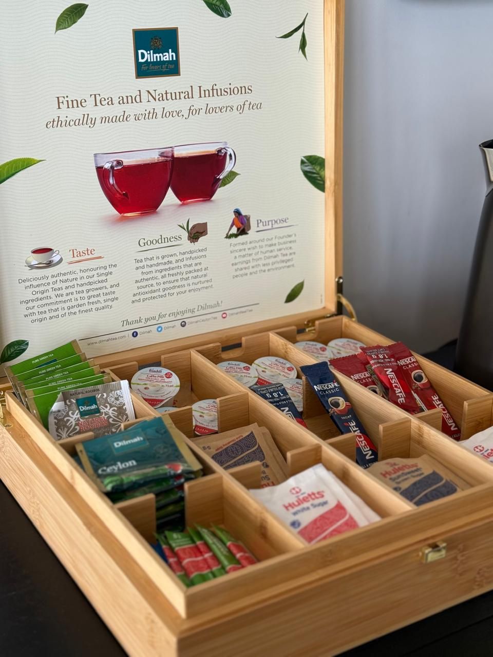 Una caja de madera con compartimentos que contienen diferentes productos de té y un cartel promocional de Dilmah.