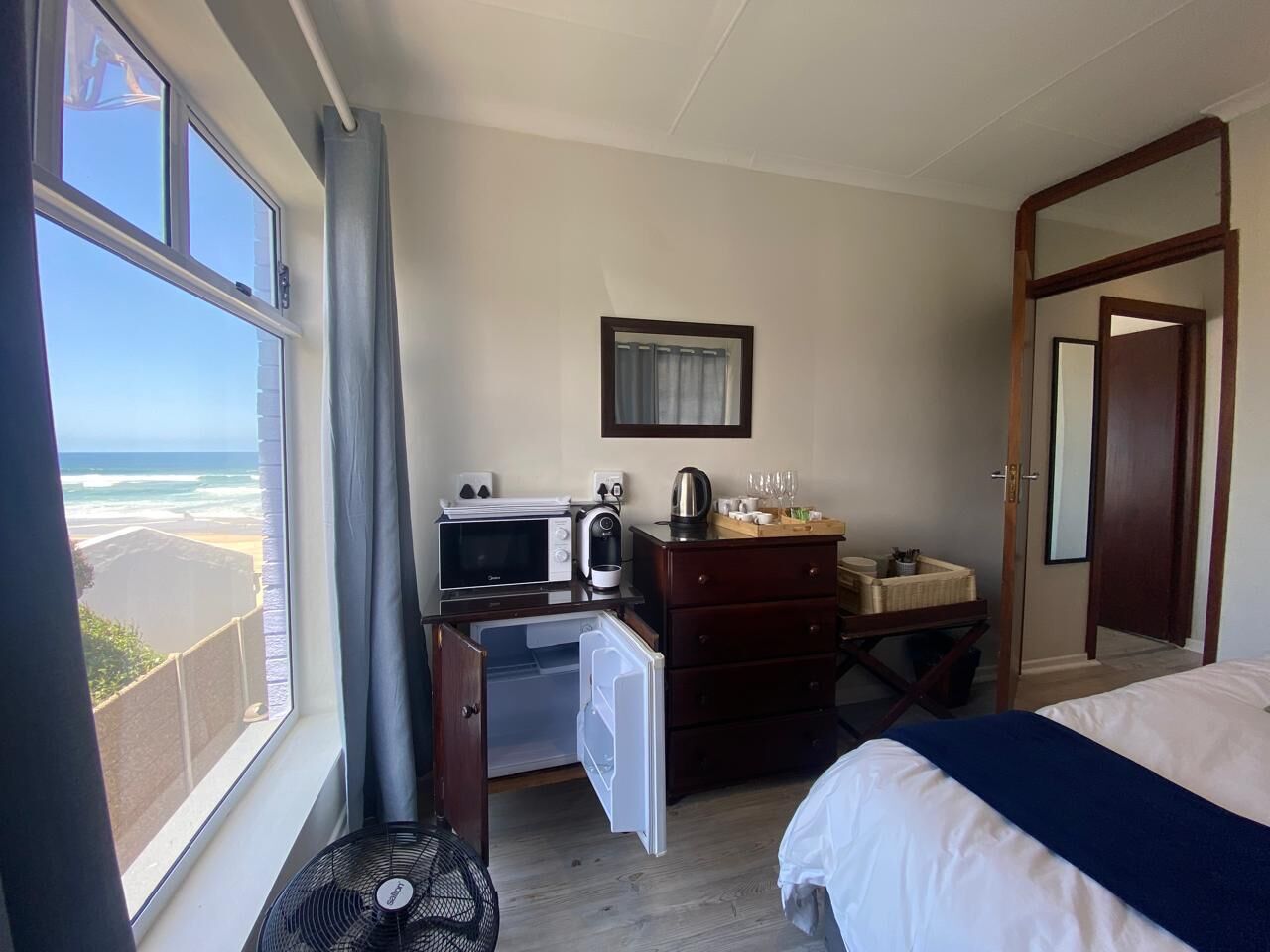 A hotel room with a bed, a desk with a microwave, a coffee maker, and a dresser. There is a window with a view of the beach.
