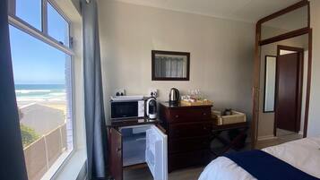 A hotel room with a bed, a desk with a microwave, a coffee maker, and a dresser. There is a window with a view of the beach.
