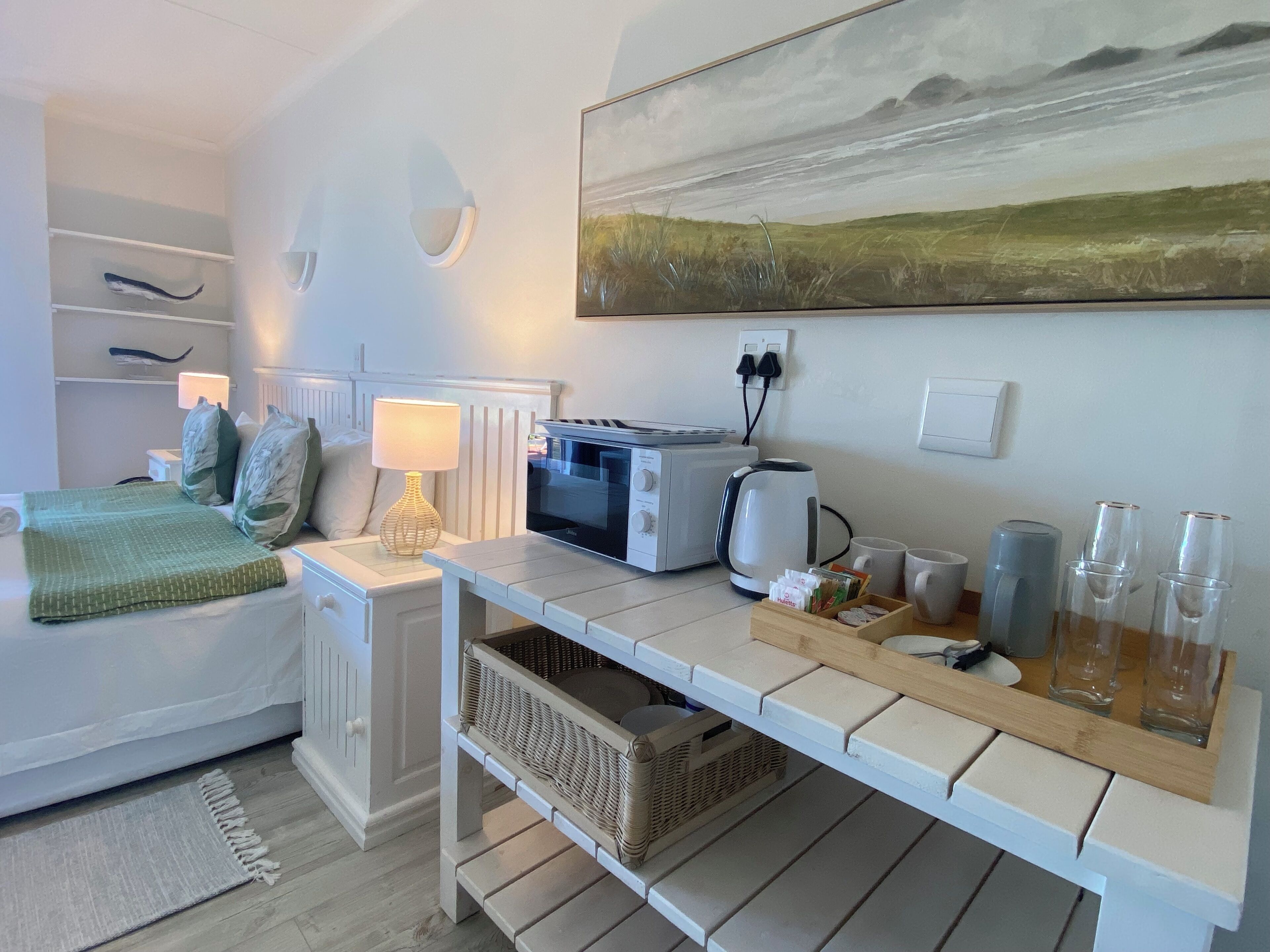 Et moderne, lyst værelse med seng, sengebord, lampe og et landskabsmaleri.