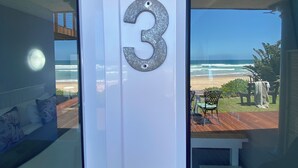 A room with a large glass door, a number '3' sign, a view of the beach, and a small outdoor seating area. - Sea Paradise (Wilderness)