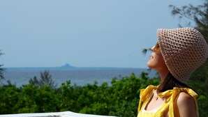 A person wearing a yellow floral dress and a beige knitted hat, standing by a concrete wall with a view of the sea and greenery. - Ocean Villa Tin Gala (Onna)
