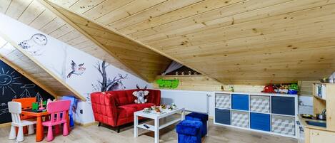 Un acogedor dormitorio en el ático con techo inclinado, un sofá rojo, una mesa blanca y una zona de juegos infantiles con juguetes y una pared con pizarra.