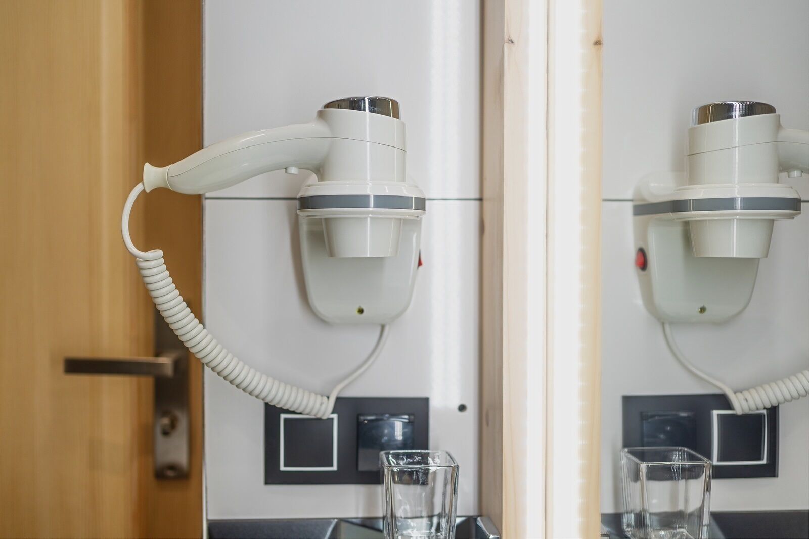 A bathroom with a hairdryer mounted on the wall, a glass of water on the counter, and a door with a handle.