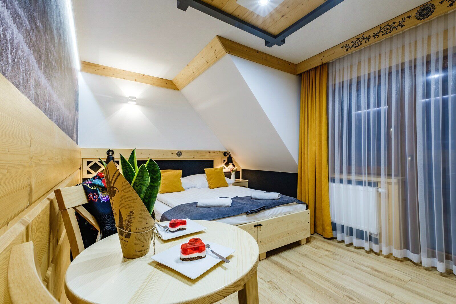 A bedroom with a sloped ceiling, a bed with yellow pillows, a bedside table with a plate of food, and a window with sheer curtains.