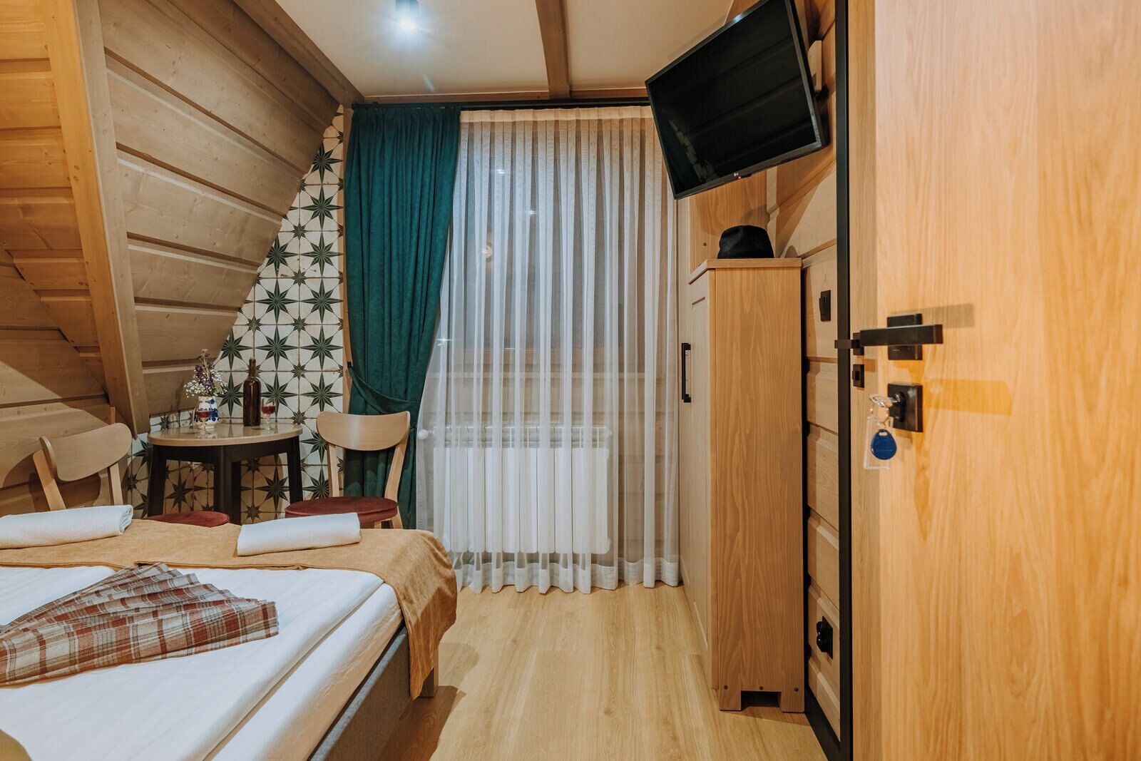 A bedroom with a bed, a small table with a vase and bottle, a chair, a TV mounted on the wall, and a wardrobe.