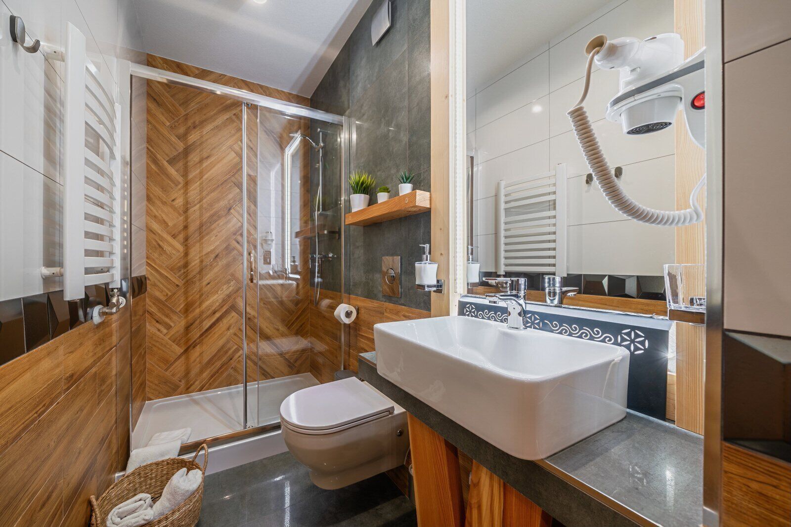 Un baño moderno con cabina de ducha de vidrio, un lavabo blanco y suelo de madera en patrón de espiga.