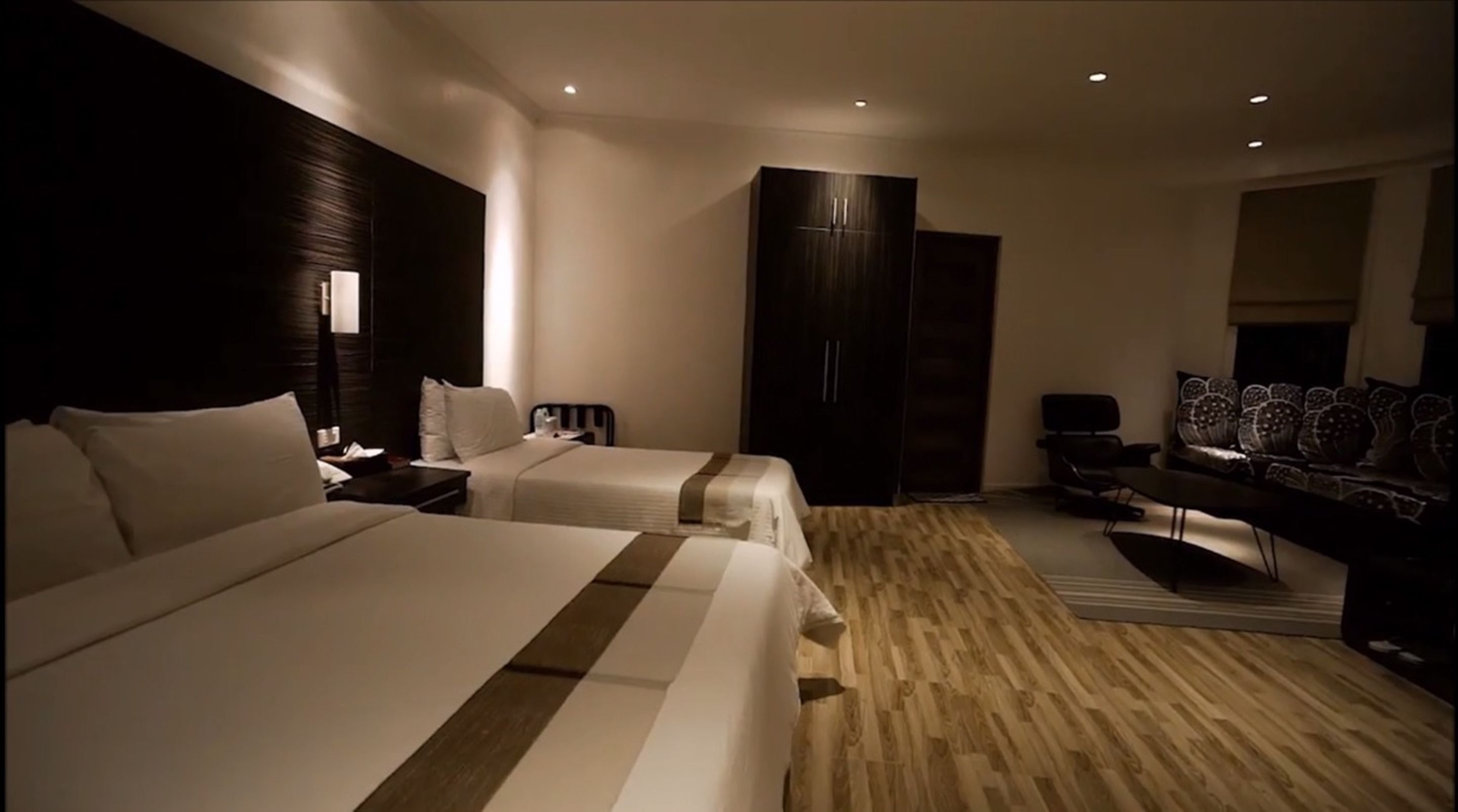Une chambre d’hôtel moderne avec deux lits, une armoire sombre, un coin salon avec un canapé et des fauteuils, et un téléviseur.