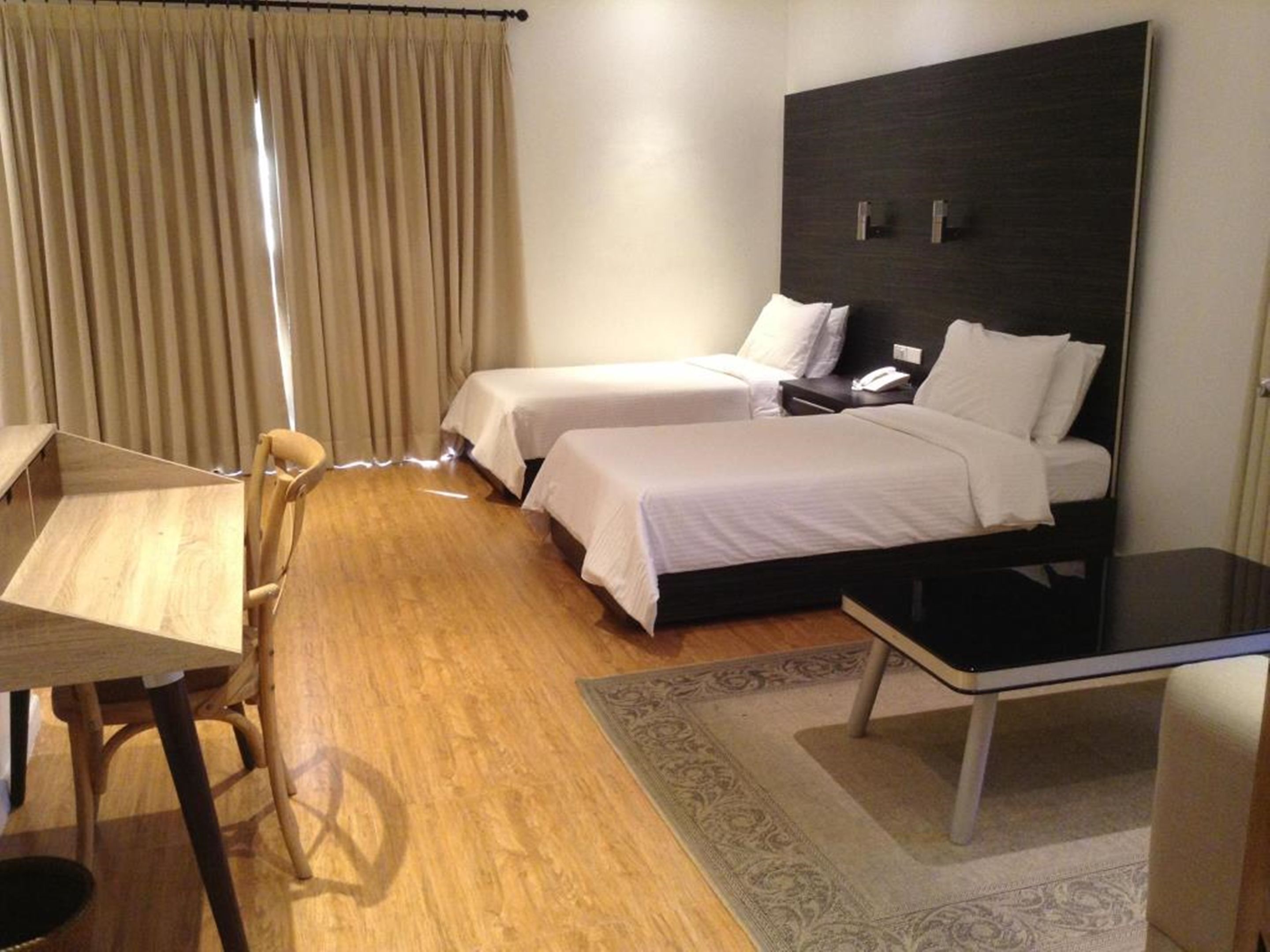 A hotel room with two single beds, a desk, a chair, a bedside table, and a large window with curtains.