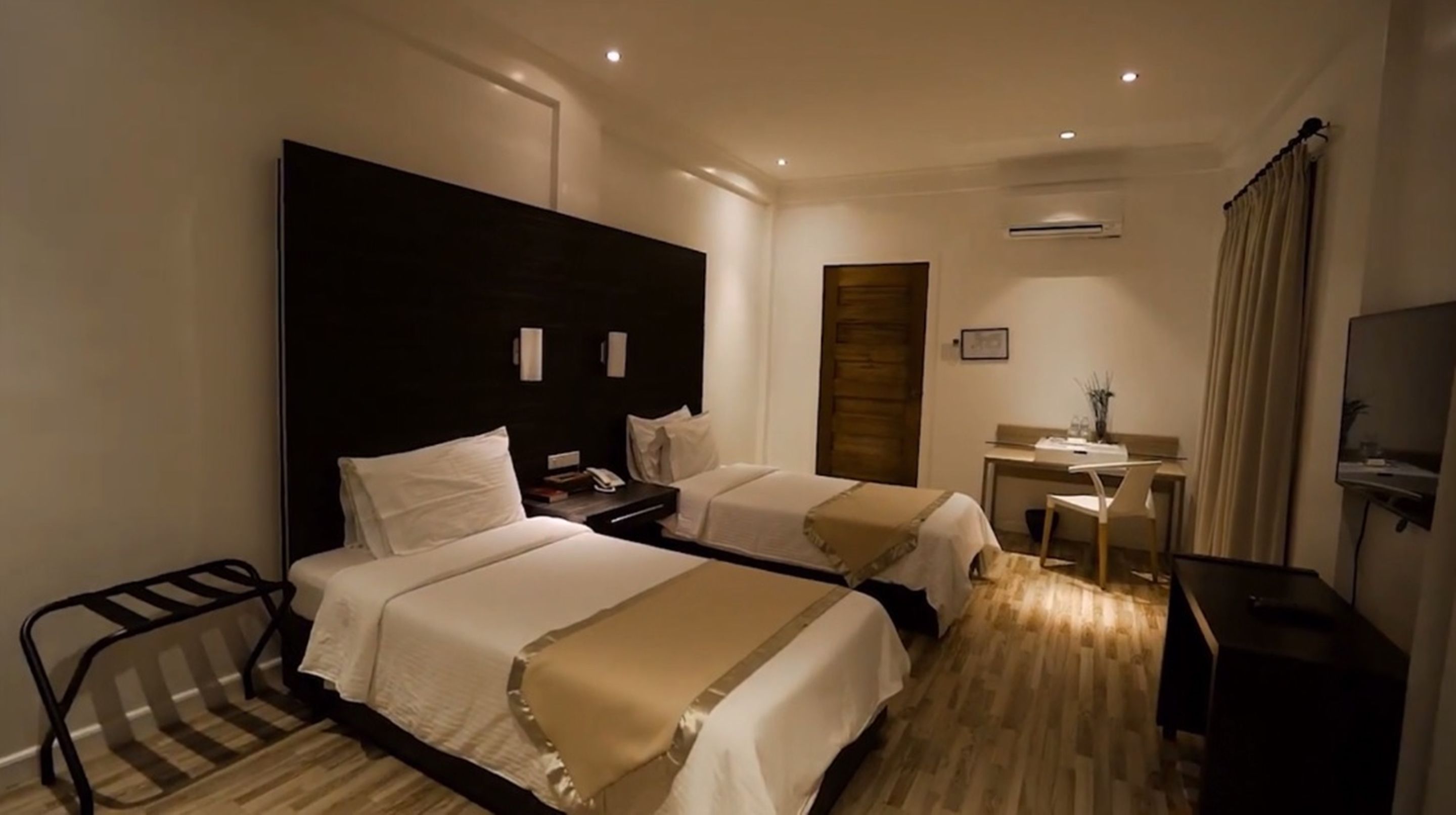 A hotel room with two beds, a desk, a chair, a TV, and an air conditioning unit.