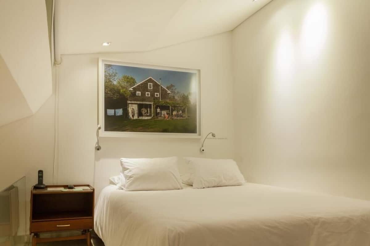 A bedroom with a bed, white pillows, a bedside table with a lamp, and a framed picture of a house on the wall.