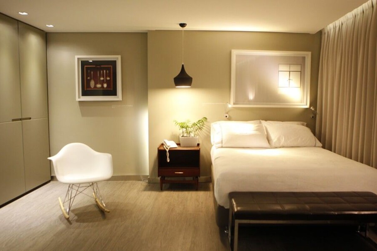 A hotel room with a bed, a chair, a bedside table with a lamp, a window with curtains, and a wall-mounted picture.