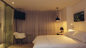 A hotel room with a bed, a chair, a desk with a lamp, a TV mounted on the wall, and a painting on the wall. - The Grace Hotel (Bogotá)