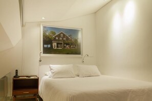 A bedroom with a bed, white pillows, a bedside table with a lamp, and a framed picture of a house on the wall. - The Grace Hotel (Bogotá)