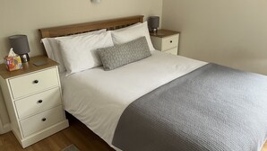 A neatly made bed with a grey blanket, two bedside tables with lamps, a wall-mounted light, and a framed picture on the wall. - Cherry Tree Cottage (Chipping Norton)