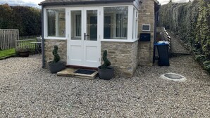 A small stone cottage with a white door and windows, a gravel driveway, and a wooden fence in the background. - Cherry Tree Cottage (Chipping Norton)
