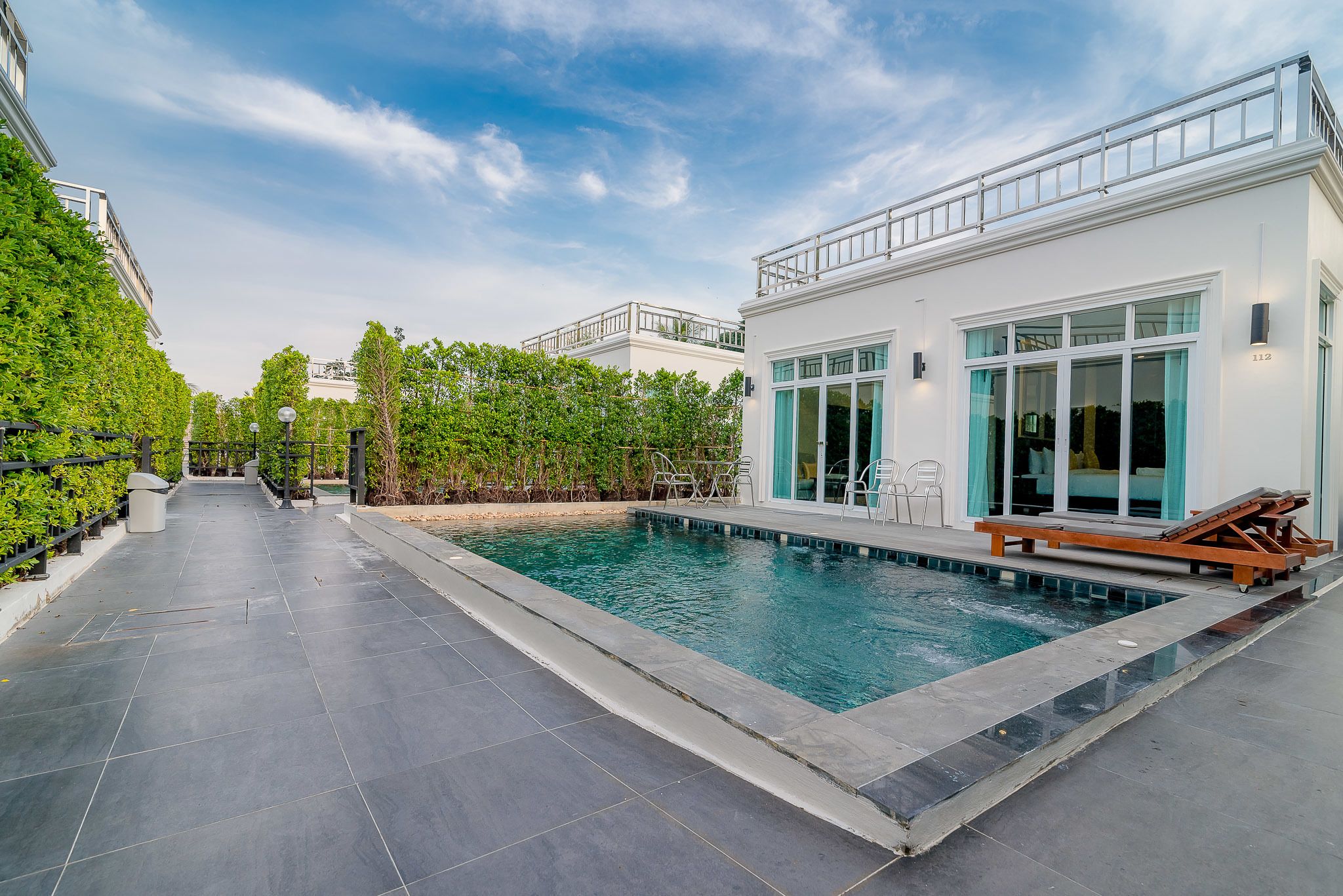 Une piscine avec une terrasse carrelée, un espace détente avec des fauteuils et un bâtiment doté de grandes fenêtres et d’un balcon.