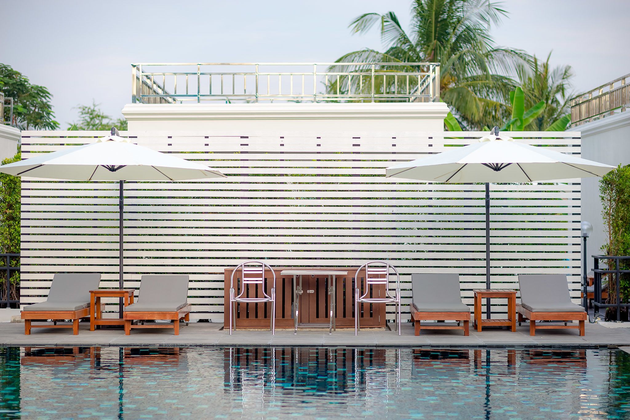 Zona de piscina con sillones de descanso y sombrillas, valla de madera y palmeras al fondo.