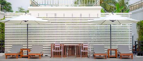 Zona de piscina con sillones de descanso y sombrillas, valla de madera y palmeras al fondo.