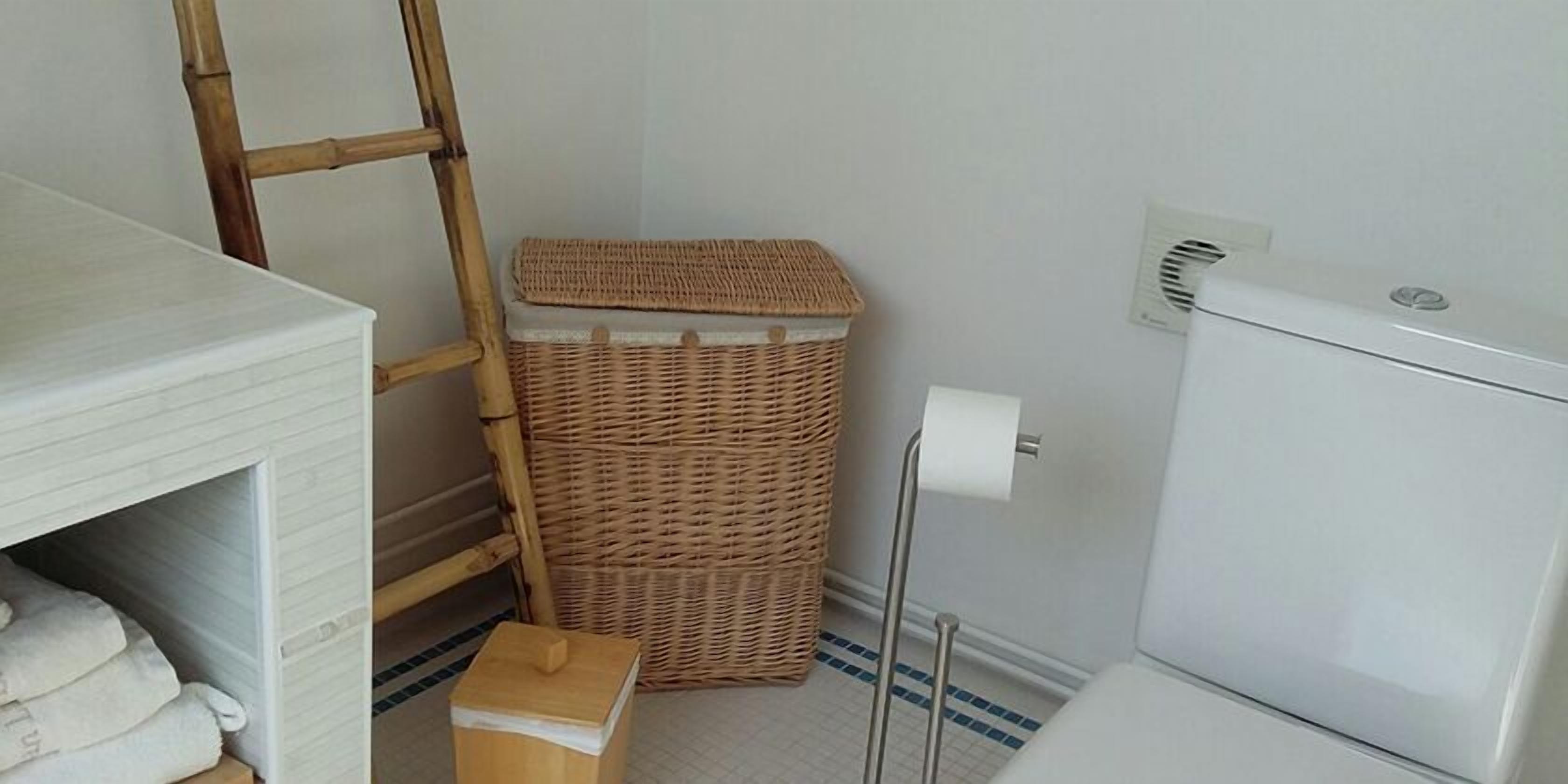 A bathroom with a white toilet, a wicker trash bin, a wooden stool, and a white towel rack.