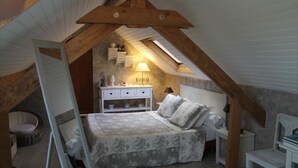 Un dormitorio con techo inclinado, una cama con colcha estampada, una mesita de noche, una lámpara y un pequeño aparador blanco.