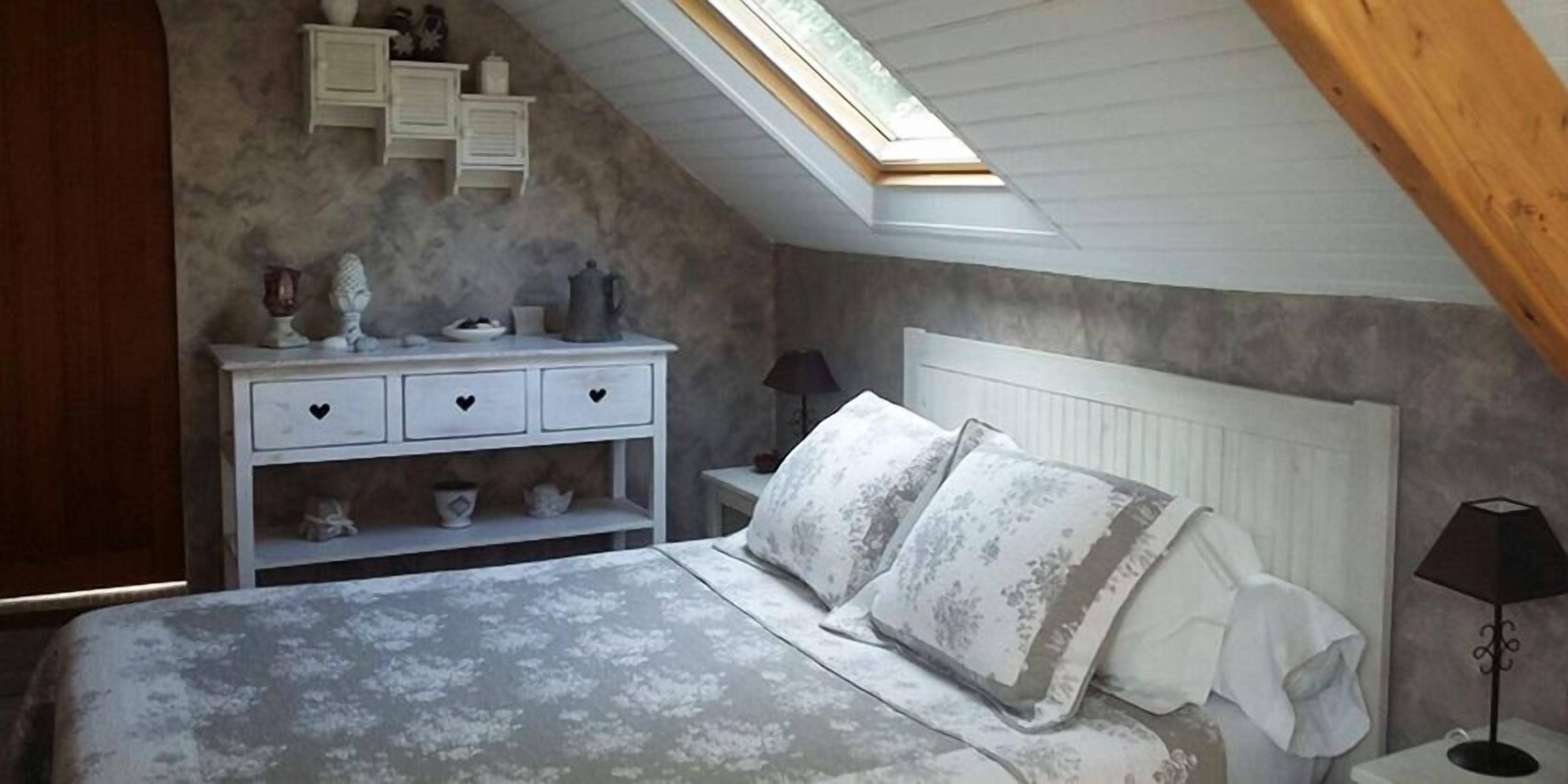 A bedroom with a sloped ceiling, a bed with patterned bedding, a nightstand with a lamp, and a dresser with decorative items.