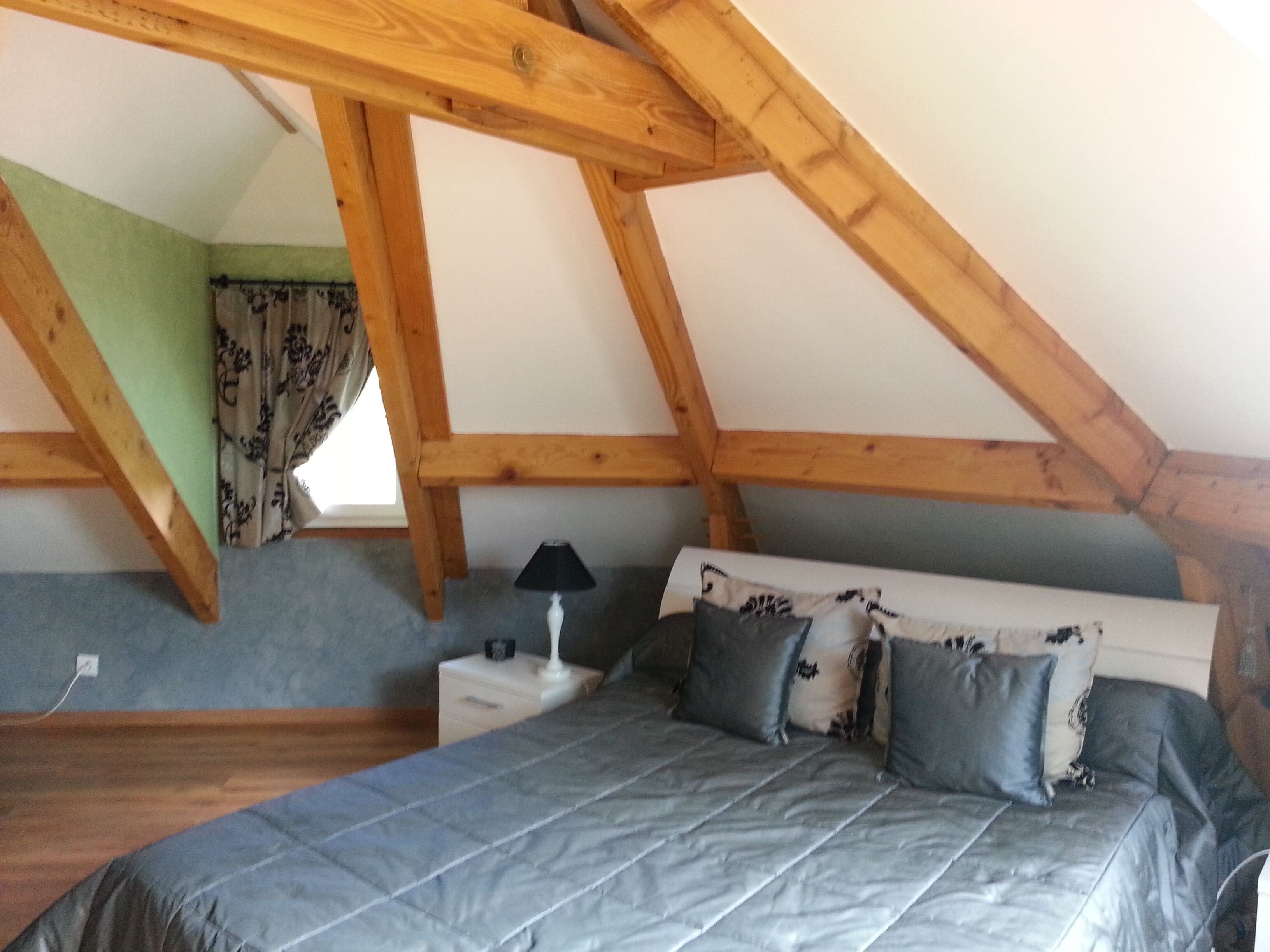A bedroom with a sloped ceiling, a bed with grey bedding, a bedside table with a lamp, and a window with curtains.