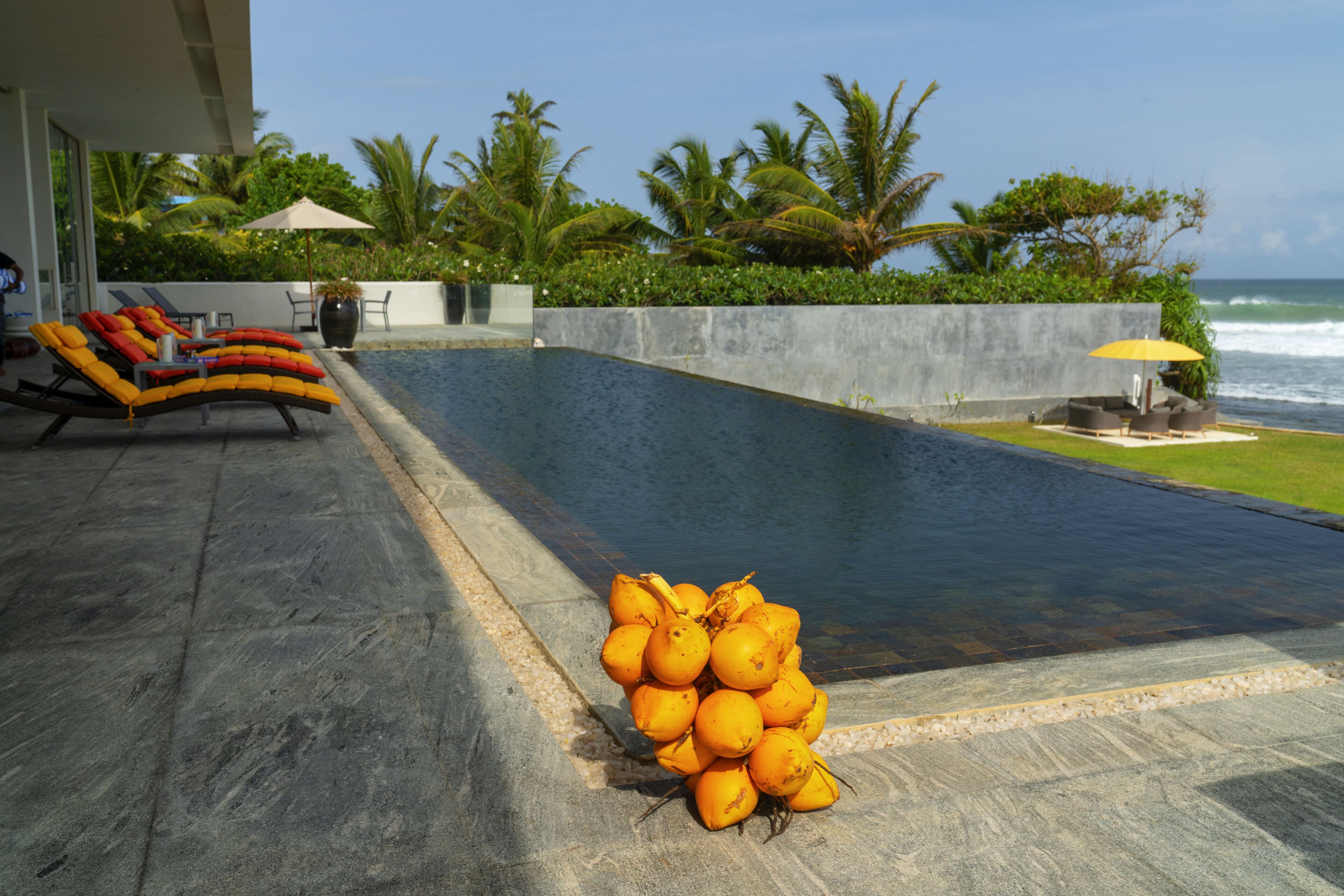 A swimming pool with a concrete edge, a lounge area with colorful cushions, and a view of the ocean.