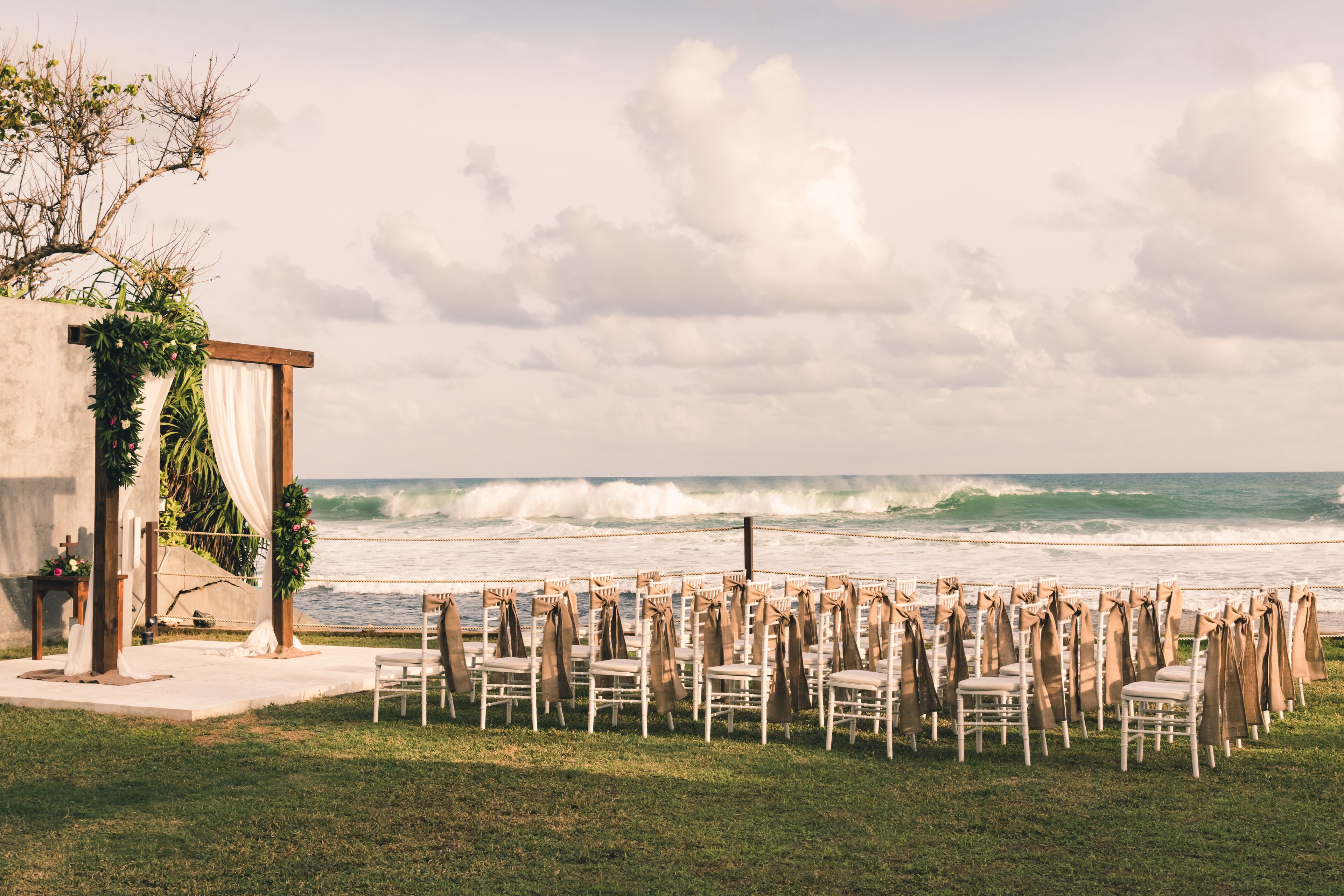 A beachside wedding setup with chairs arranged in rows facing the ocean, a small altar with draped fabric, and a backdrop of waves.