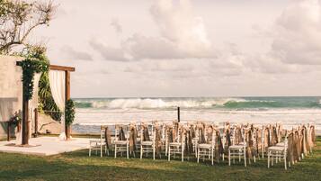 A beachside wedding setup with chairs arranged in rows facing the ocean, a small altar with draped fabric, and a backdrop of waves.