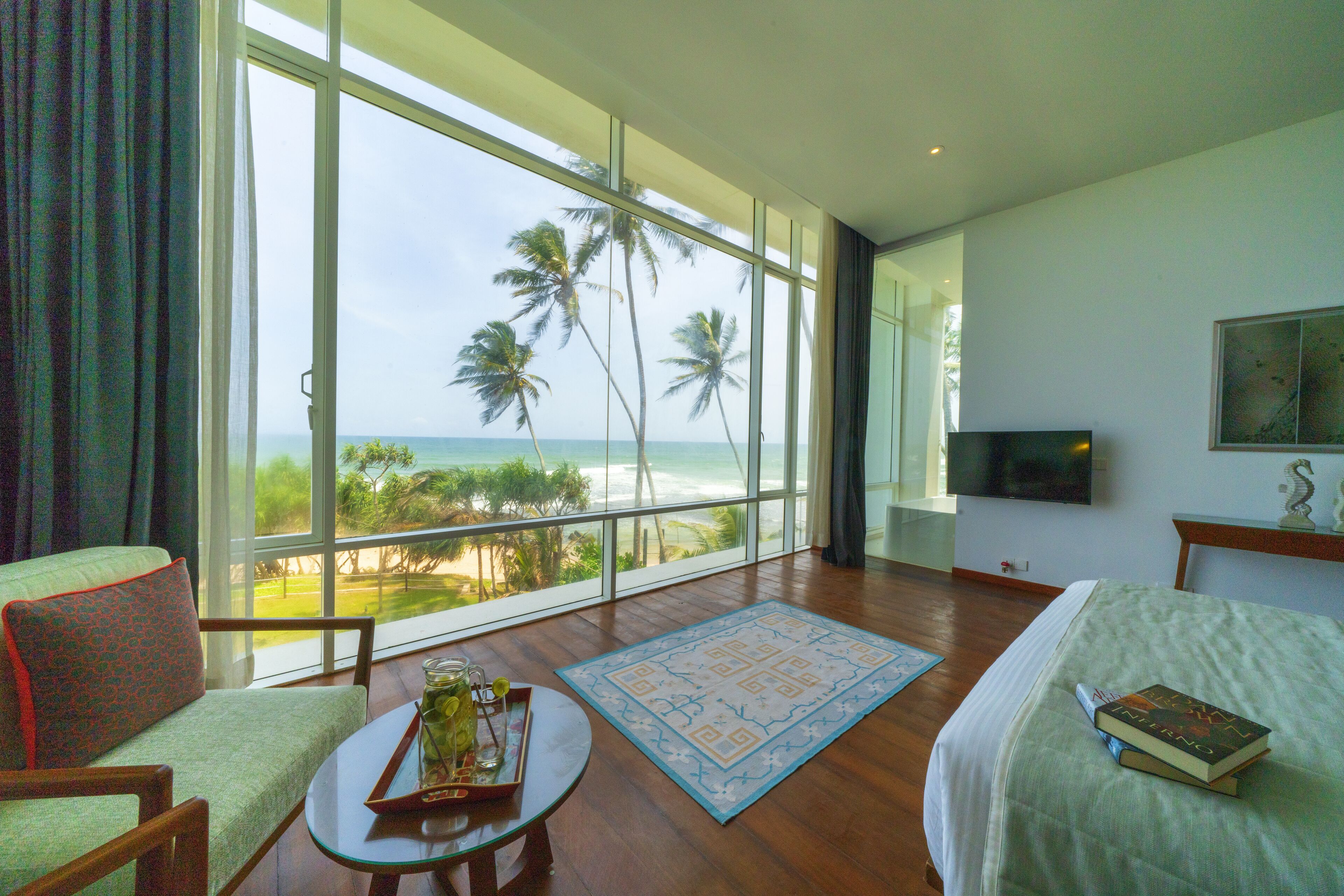 A hotel room with a large window offering a view of palm trees and the ocean. There is a bed, a TV mounted on the wall, and a small table with a tray and glass.