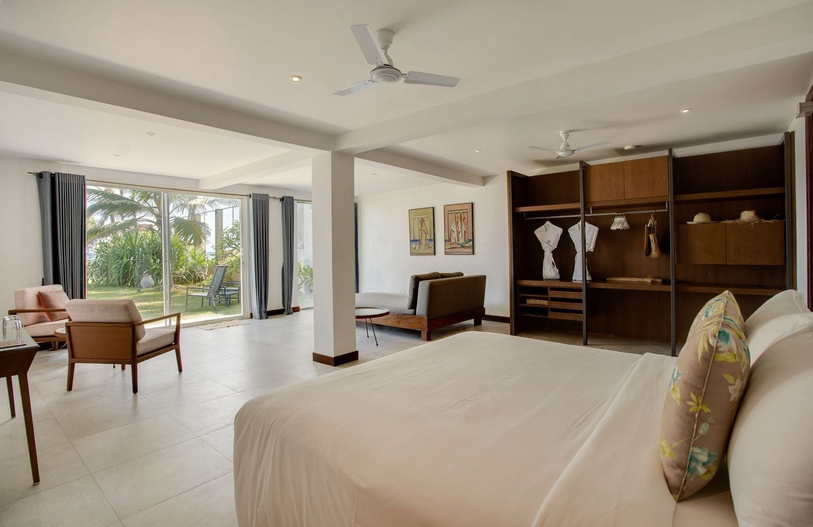 A spacious bedroom with a large bed, a seating area with a sofa and chairs, a walk-in closet, and a view of the outdoors through large windows.