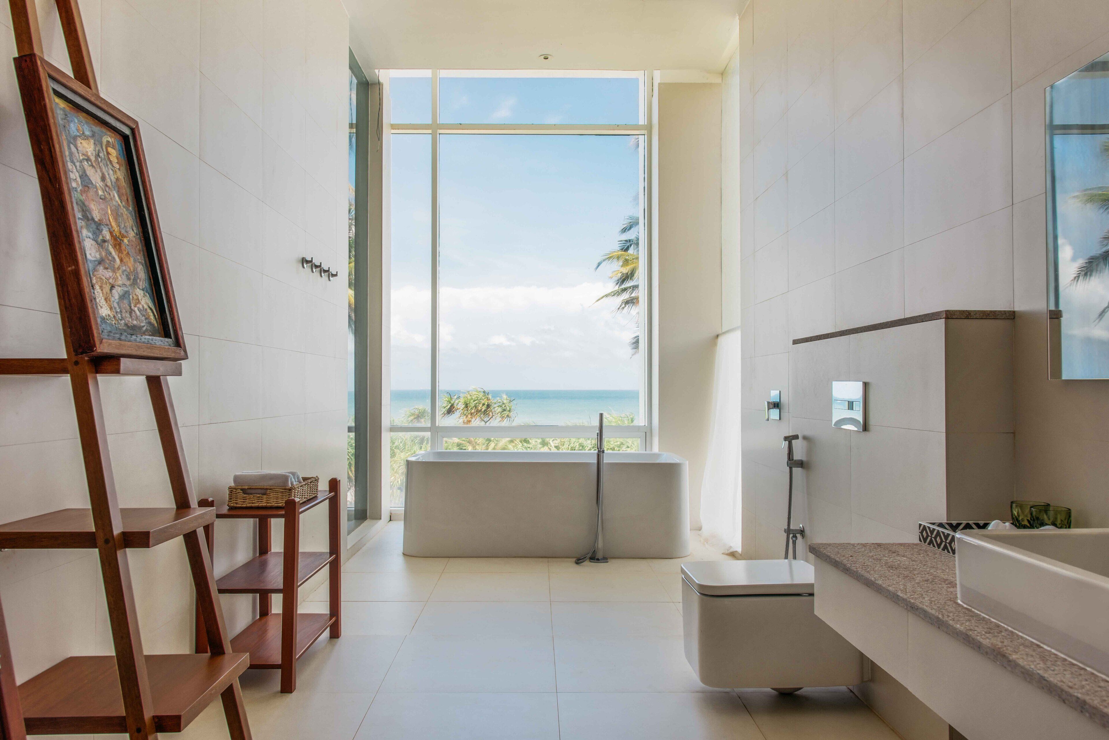 A modern bathroom with a bathtub, a shower, and a large window offering a view of the beach.