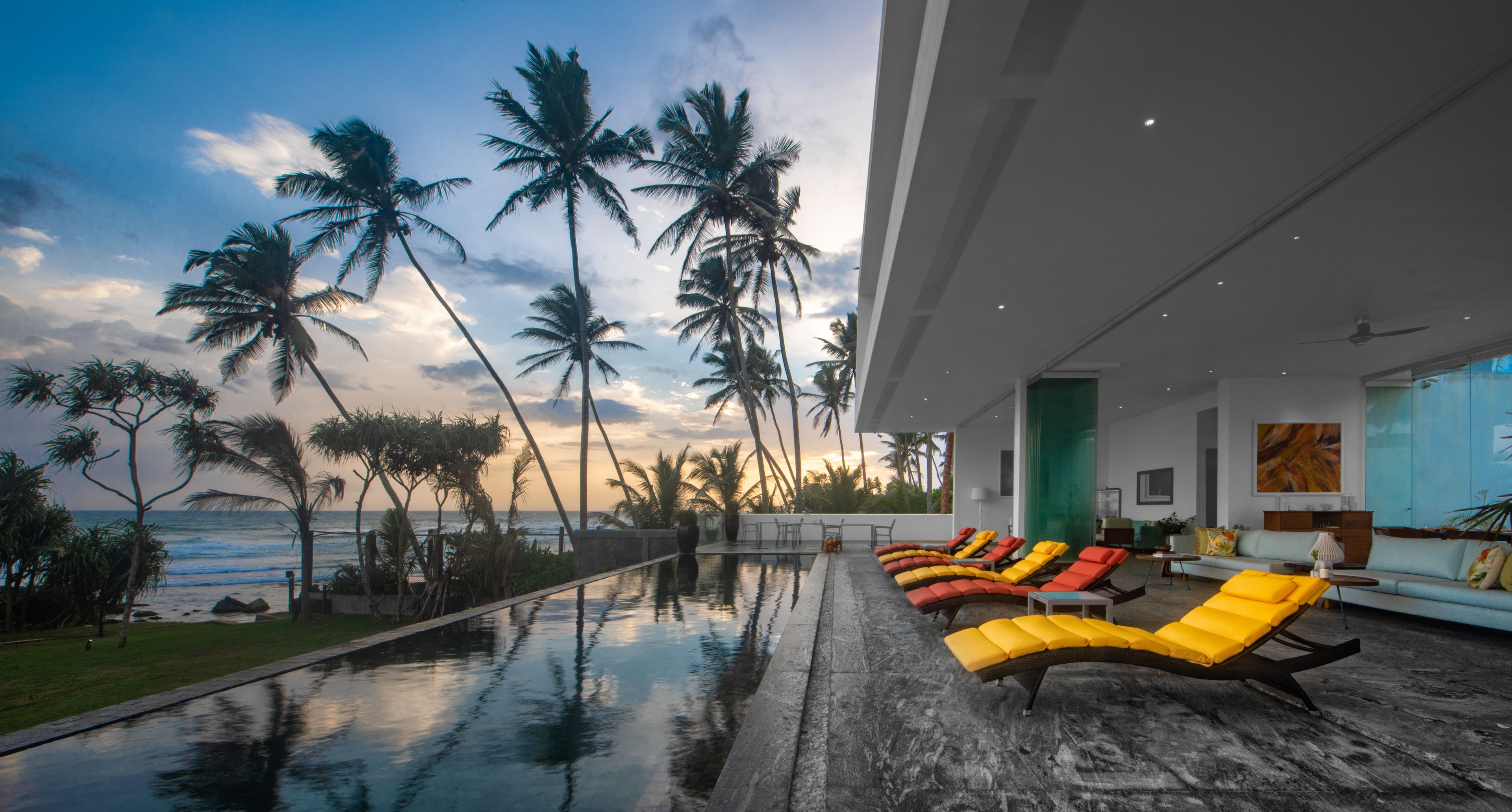 A modern outdoor lounge area with a pool, sun loungers, and a view of the ocean and palm trees.