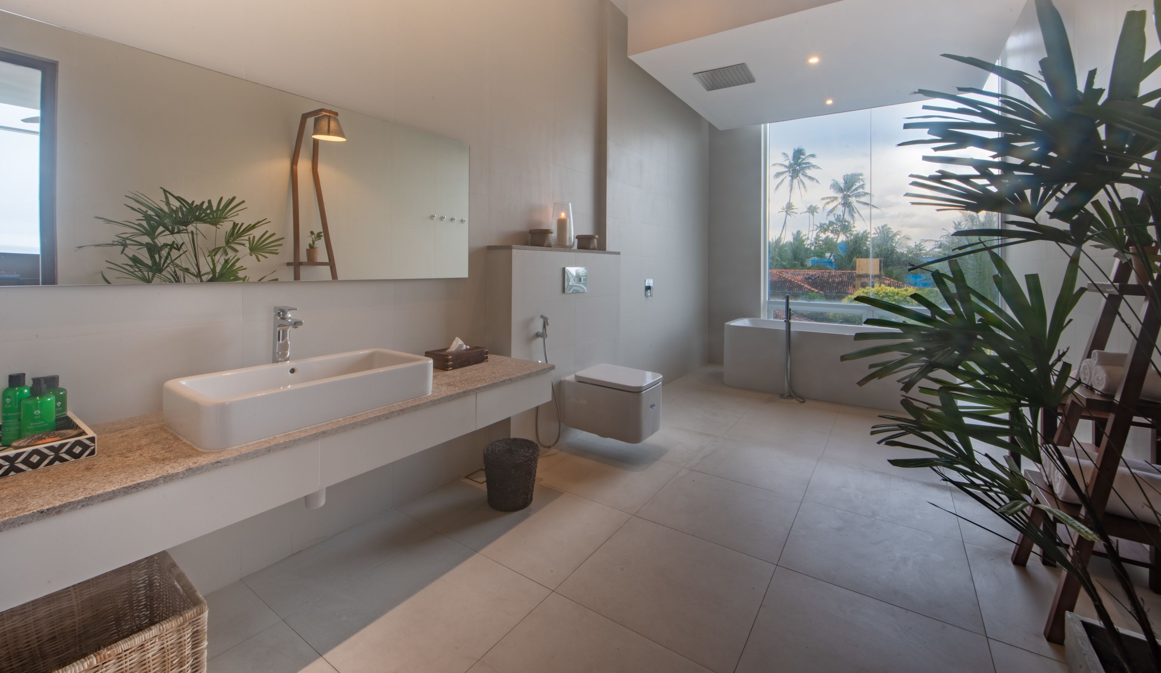Un baño moderno con un lavamanos grande, inodoro y bañera. Hay un espejo sobre el lavamanos, una lámpara de pie y varias plantas.