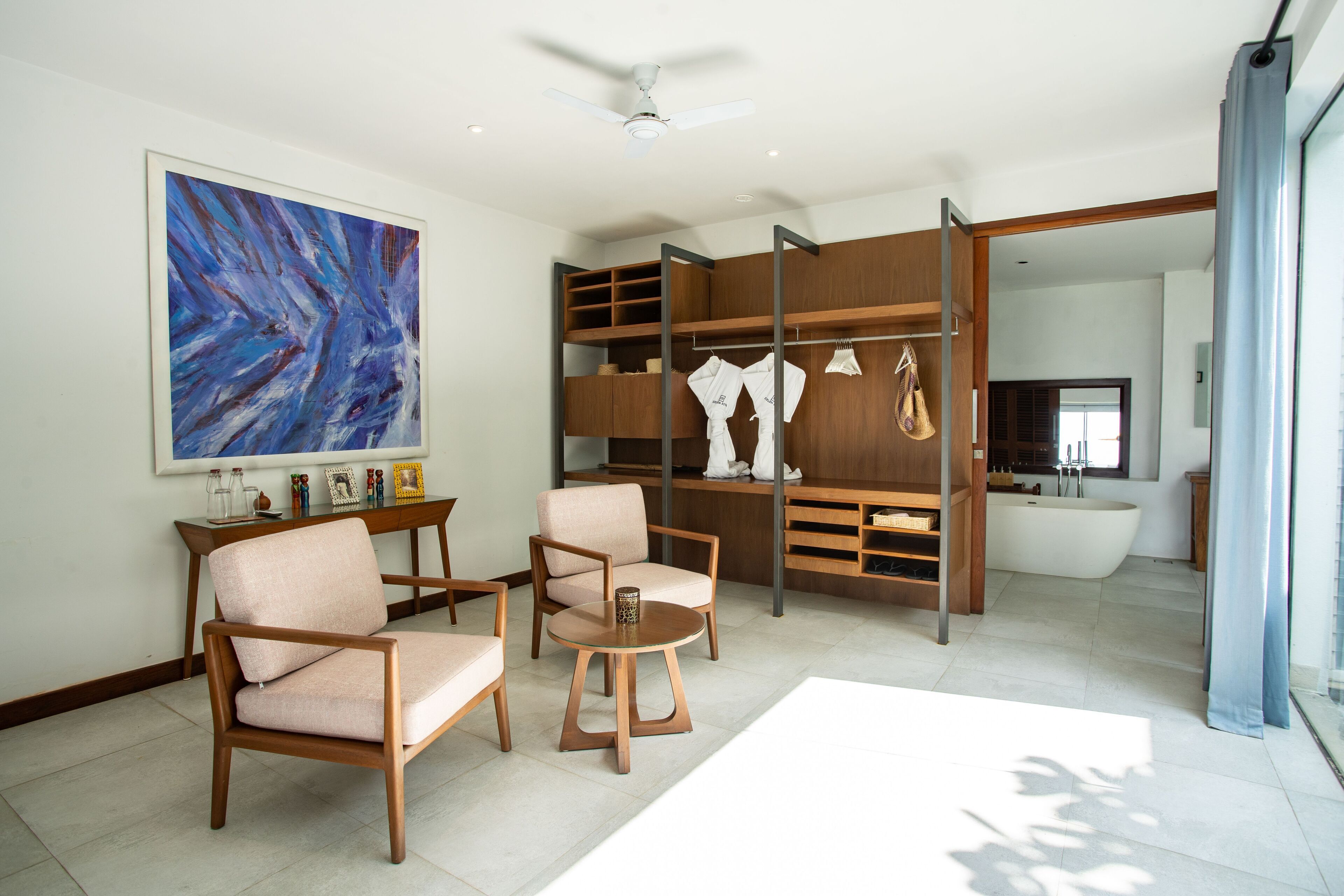 Una sala de estar moderna con un cuadro abstracto grande, dos sillas de madera, una mesita redonda y una estantería empotrada con ropa y objetos decorativos.