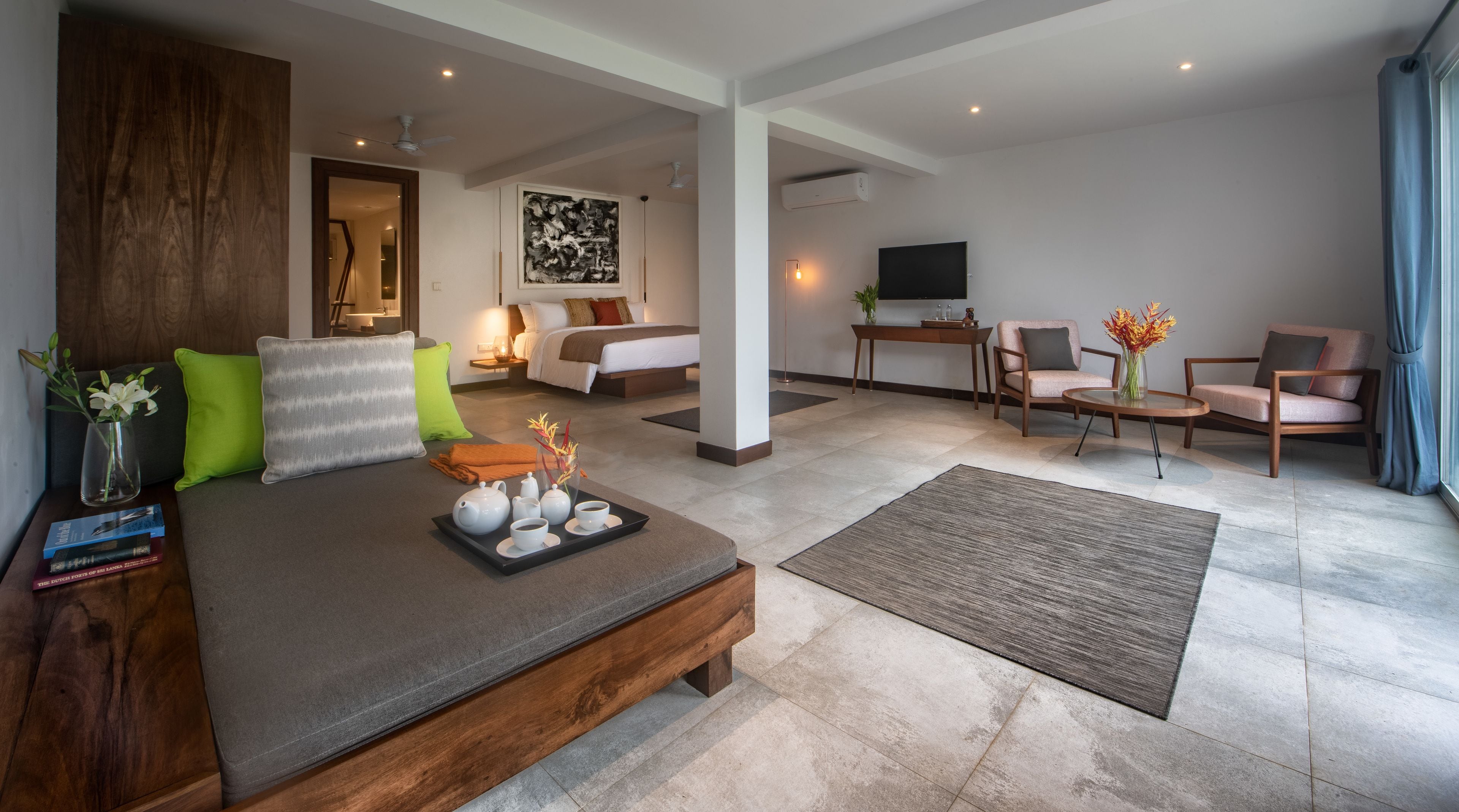 A modern hotel room with a large bed, a desk, two chairs, a TV, and a painting on the wall.