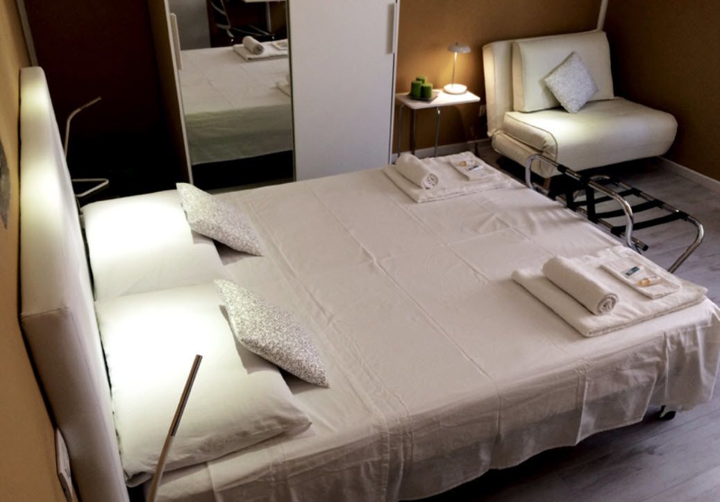 Una camera d'albergo con un letto grande, una sedia, un tavolino con una lampada e uno specchio.