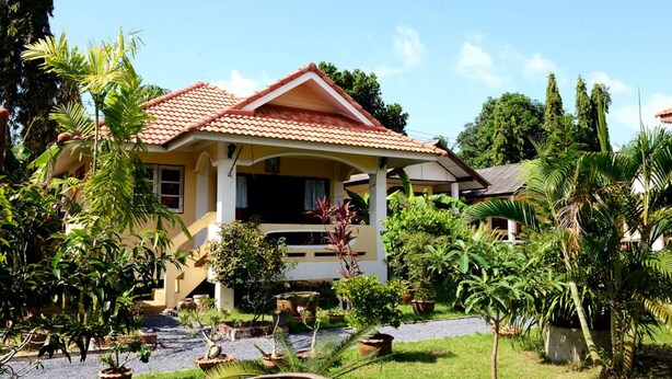 Une maison à deux étages, avec un toit recouvert de tuiles rouges, une véranda et un jardin où poussent différentes plantes, ainsi qu’un petit étang.