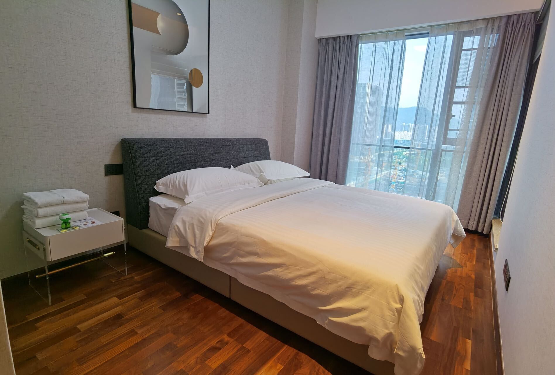 Habitación de hotel con cama, mesita de noche con artículos de aseo, ventana grande con cortinas y espejo adosado a la pared.