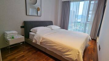 Habitación de hotel con cama, mesita de noche con artículos de aseo, ventana grande con cortinas y espejo adosado a la pared.