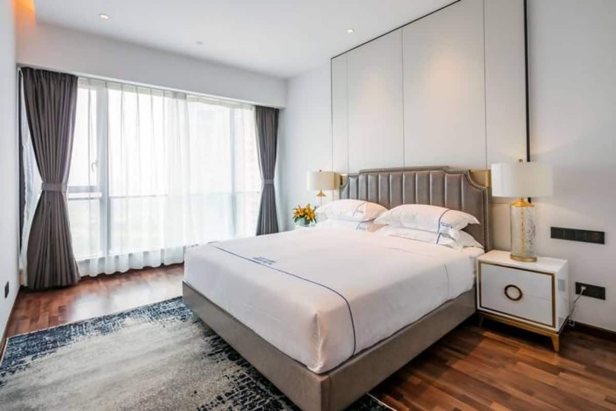 Ein modernes Hotelzimmer mit einem großen Bett, einem Nachttisch mit Lampen, Blick auf die Stadt durch große Fenster und Holzboden.