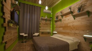 Una habitación con cama, dos sillas, televisor y estanterías con plantas.