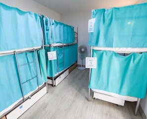 Una habitación con literas, cada una cubierta con sábanas azul turquesa, y un ventilador en el suelo.