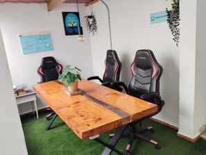 Un salón de conferencias con una mesa de madera, sillas tipo gamer negras, una planta en maceta y adornos en la pared.