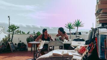 Two people sitting at a table on a rooftop terrace with potted plants and a view of the ocean.