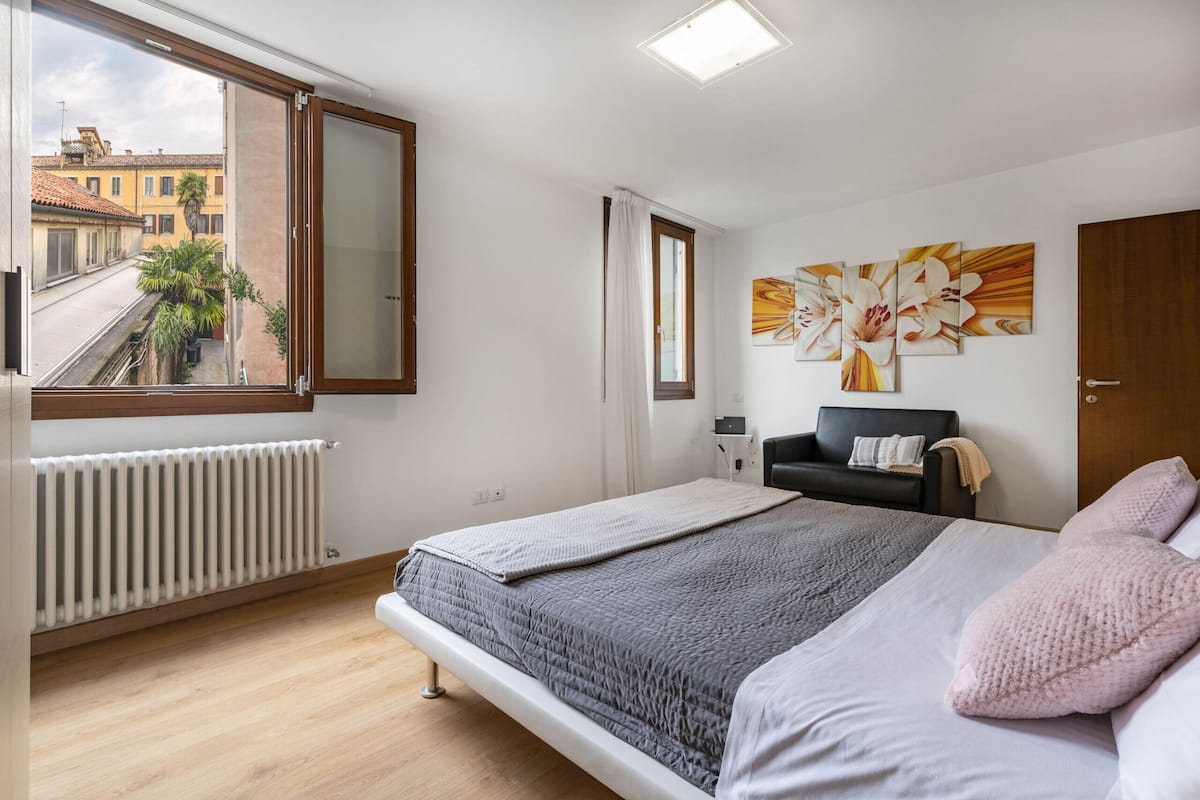 A bedroom with a large window, a radiator, a bed with a grey quilt, a black chair, and a wall with a floral painting.