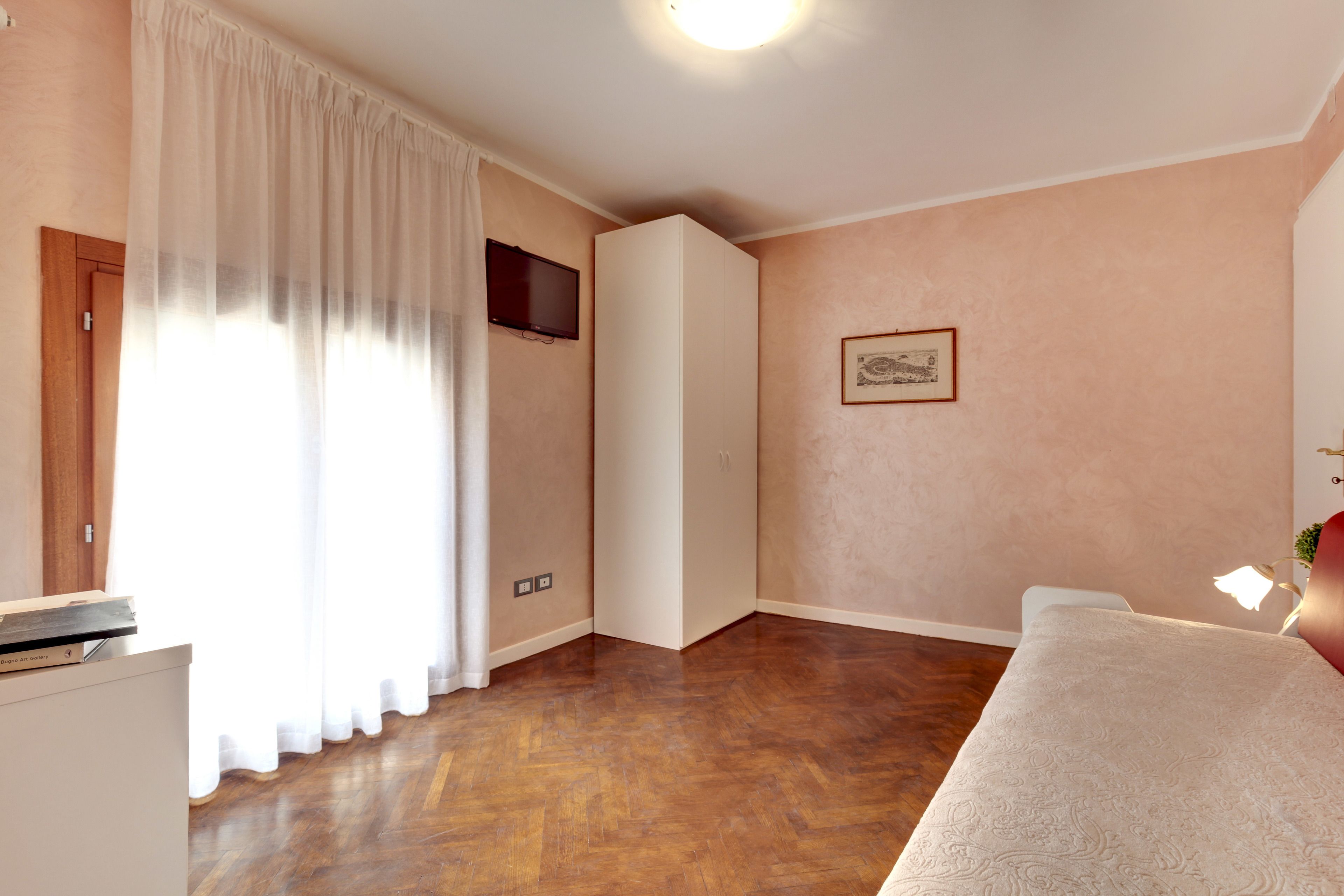 A room with a wooden floor, a white bed, a television mounted on the wall, and a door.