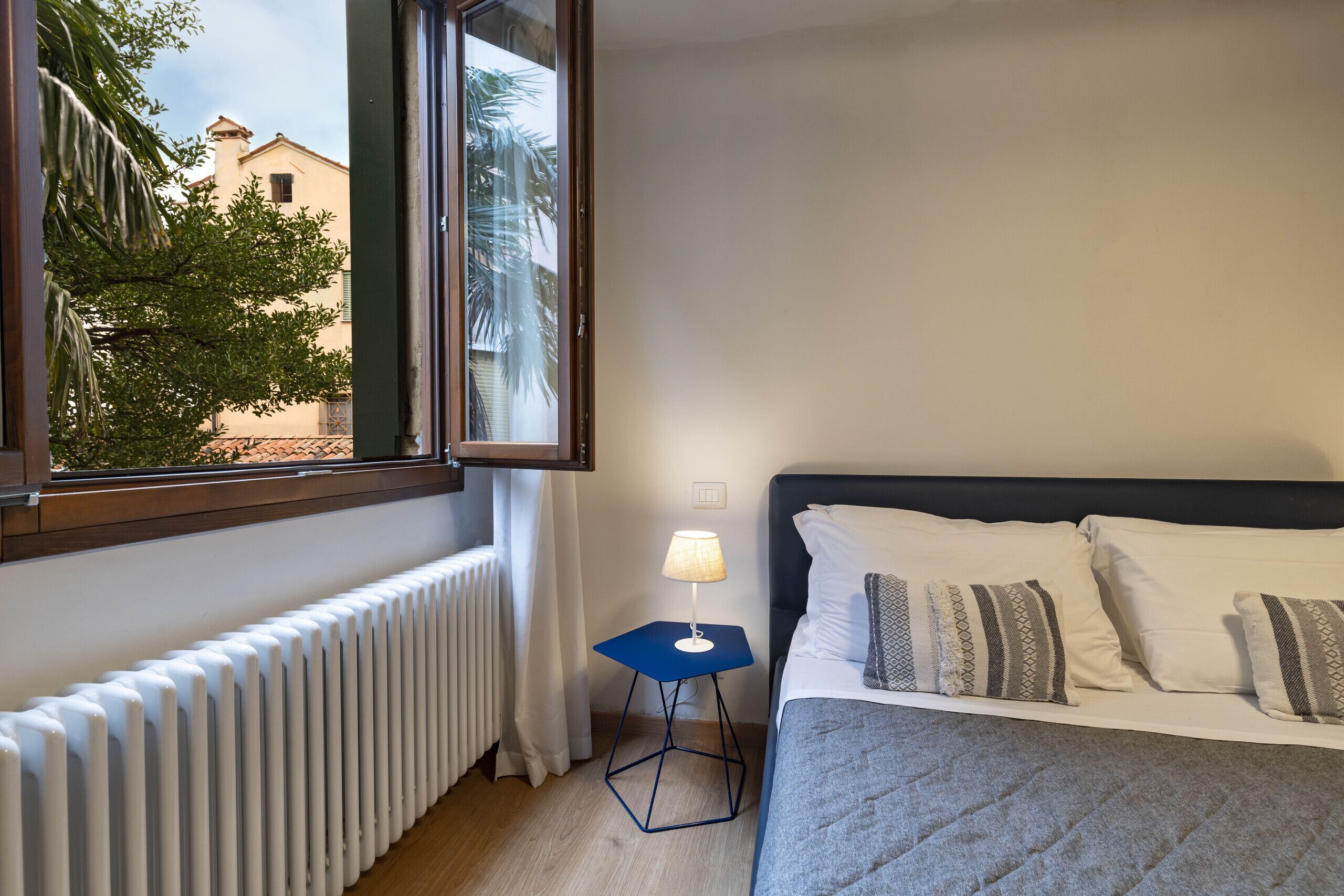 A hotel room with a bed, a window with a view of buildings and trees, a radiator, and a small table with a lamp.